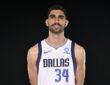 Dallas Mavericks forward Santi Aldama smiles in a white No. 34 Dallas jersey against a black backdrop.