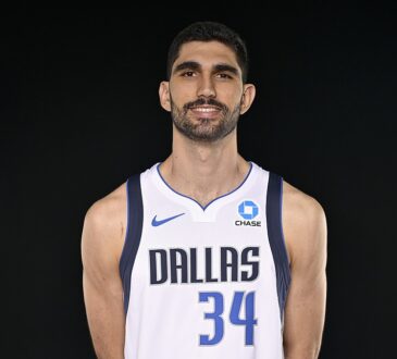 Dallas Mavericks forward Santi Aldama smiles in a white No. 34 Dallas jersey against a black backdrop.