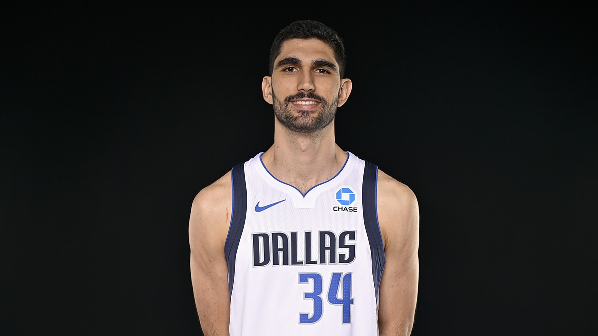 Dallas Mavericks forward Santi Aldama smiles in a white No. 34 Dallas jersey against a black backdrop.