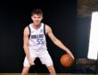 Dallas Mavericks guard Sergio de Larrea smiles and dribbles a basketball in a white home uniform in front of a black backdrop.