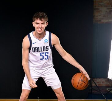 Dallas Mavericks guard Sergio de Larrea smiles and dribbles a basketball in a white home uniform in front of a black backdrop.