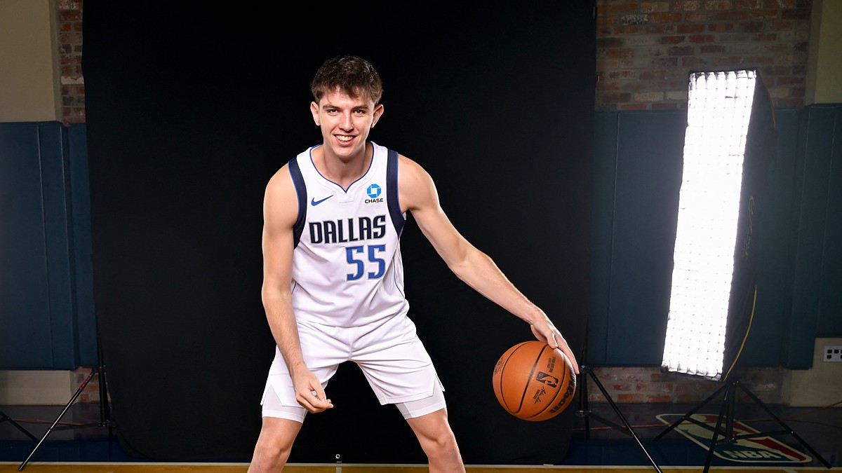 Dallas Mavericks guard Sergio de Larrea smiles and dribbles a basketball in a white home uniform in front of a black backdrop.