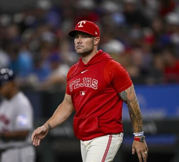 Texas Rangers manager Skip Schumaker walks across the infield in a red Tejas hooded top.