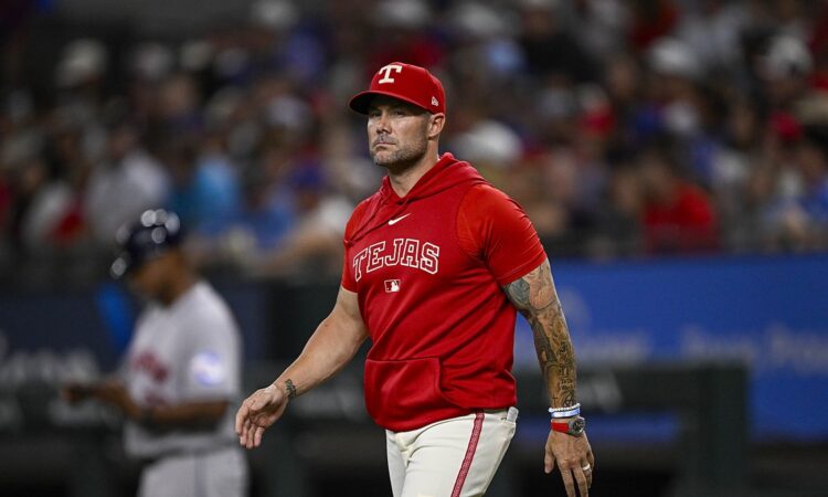 Texas Rangers manager Skip Schumaker walks across the infield in a red Tejas hooded top.