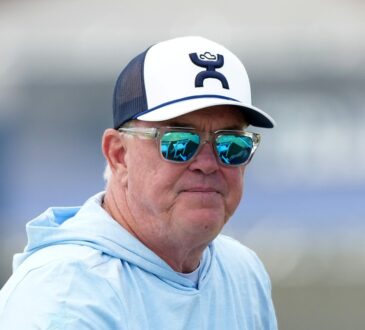 Cowboys chief operating officer Stephen Jones watches training camp in sunglasses, a ball cap, and a light blue hoodie.