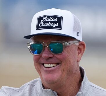 Cowboys chief operating officer Stephen Jones smiles at training camp in a ball cap and sunglasses.
