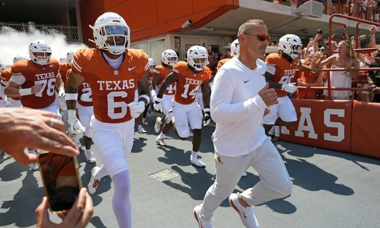 Texas Longhorns head coach Steve Sarkisian leads his players onto the field as fans cheer from the stands.