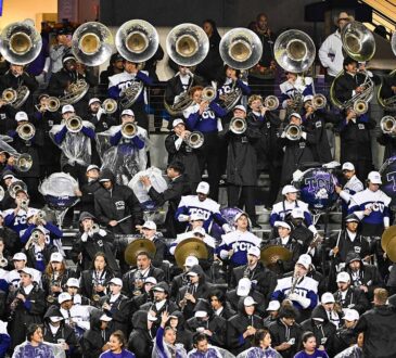 TCU Horned Frogs marching band members in rain gear play sousaphones, drums, and cymbals in the stands.