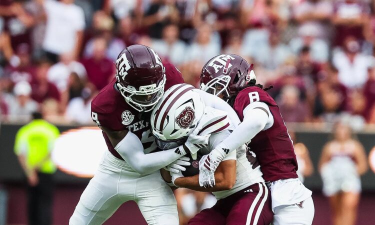 Two Texas A&M defenders wrap up a Missouri State ball carrier who holds the ball against his chest.