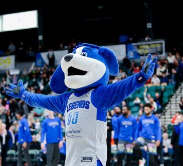Texas Legends mascot Dunker spreads his arms on the court as players in warmup shirts stand behind him.