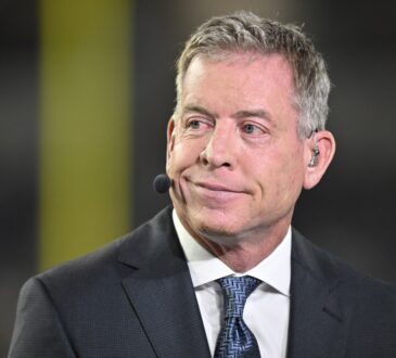 Television commentator Troy Aikman speaks on camera in a suit and earpiece before the Cowboys and Cardinals game.