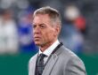 Troy Aikman stands on the field in a gray suit during pregame warmups at MetLife Stadium.