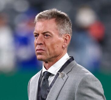 Troy Aikman stands on the field in a gray suit during pregame warmups at MetLife Stadium.