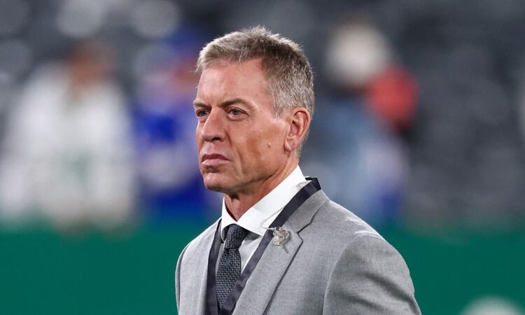 Troy Aikman stands on the field in a gray suit during pregame warmups at MetLife Stadium.