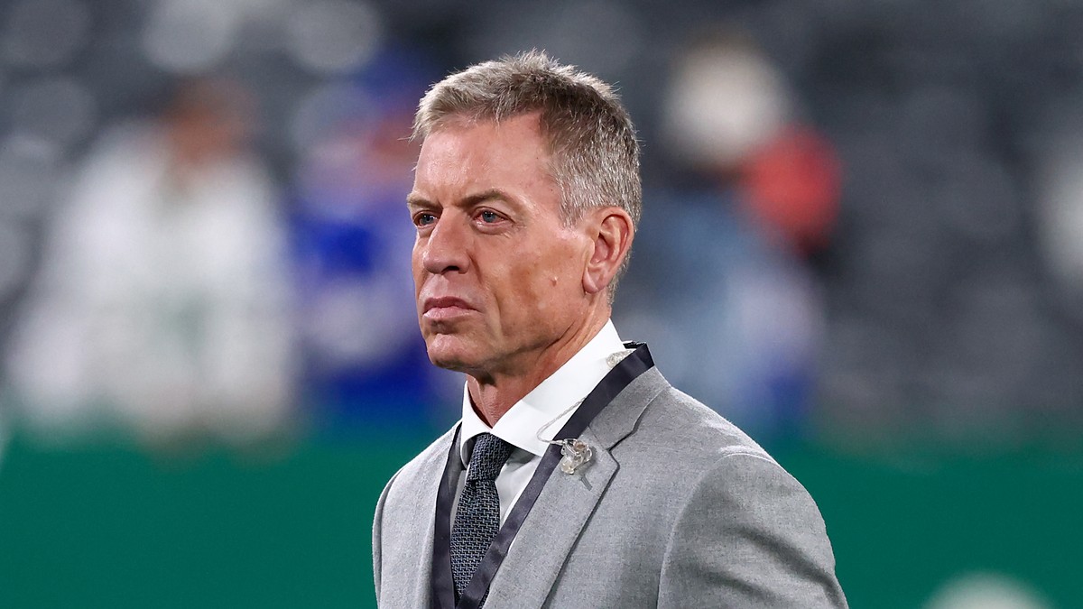 Troy Aikman stands on the field in a gray suit during pregame warmups at MetLife Stadium.