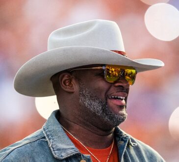 Former Texas Longhorns quarterback Vince Young smiles in a cowboy hat and sunglasses on the field during a game.