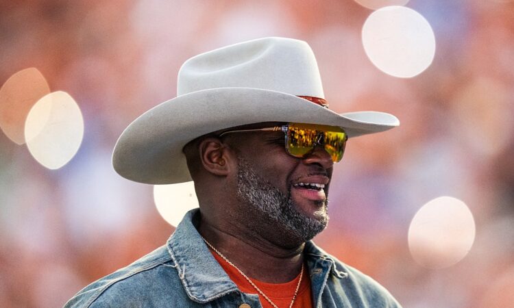 Former Texas Longhorns quarterback Vince Young smiles in a cowboy hat and sunglasses on the field during a game.