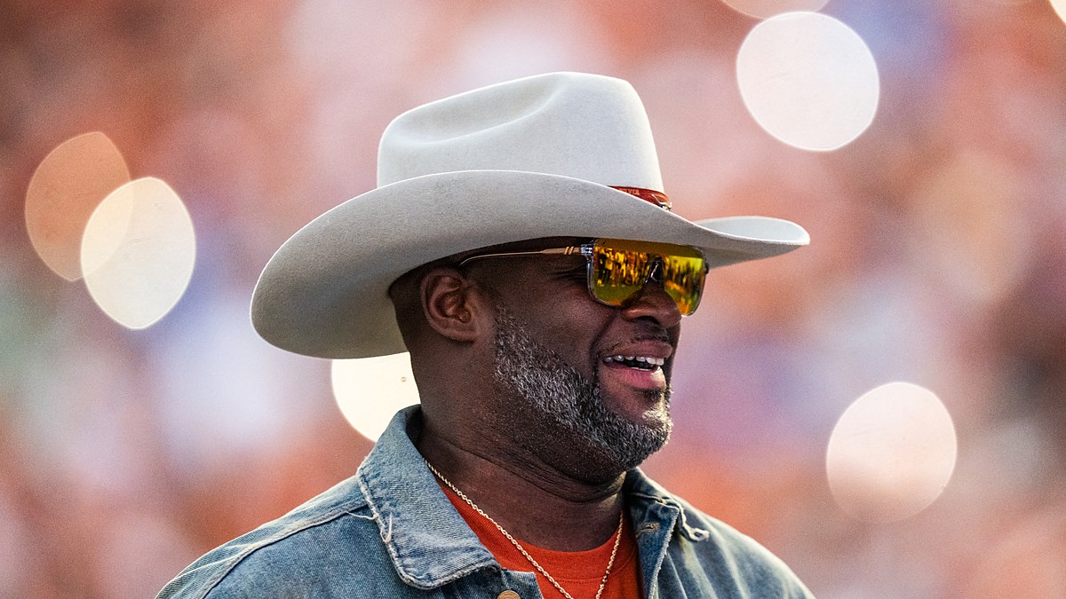 Former Texas Longhorns quarterback Vince Young smiles in a cowboy hat and sunglasses on the field during a game.
