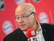 NHL deputy commissioner William Daly, in rimless glasses and a dark blazer, speaks into a Bayern Munich microphone.