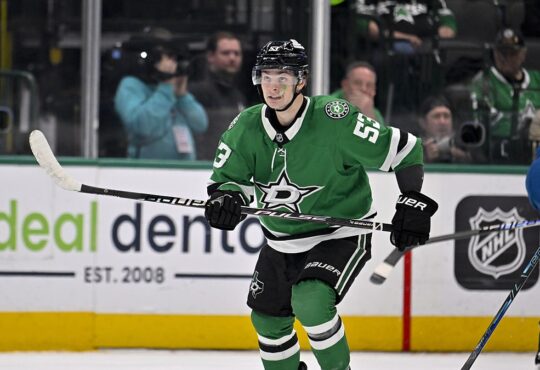 Dallas Stars center Wyatt Johnston skates with his stick across his body as Colorado Avalanche players look on.