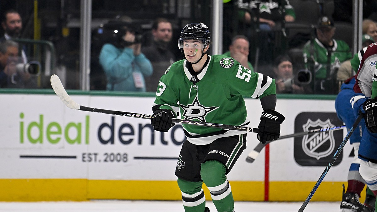 Dallas Stars center Wyatt Johnston skates with his stick across his body as Colorado Avalanche players look on.