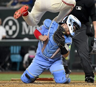 Toronto Blue Jays catcher Alejandro Kirk braces on his knees as Texas Rangers left fielder Wyatt Langford leaps over him at the plate.