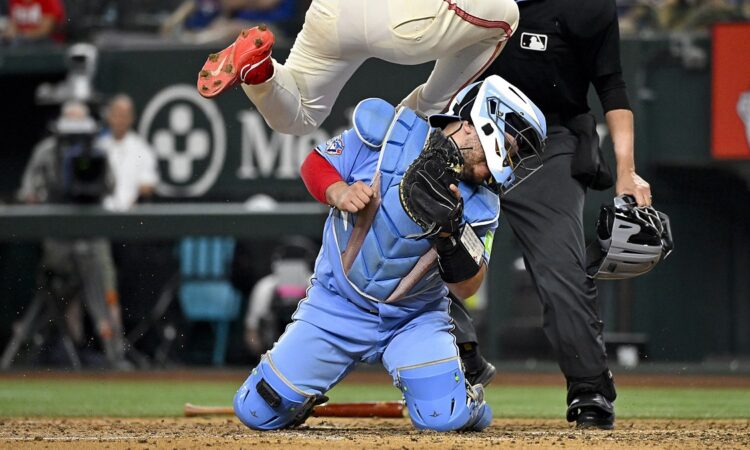 Toronto Blue Jays catcher Alejandro Kirk braces on his knees as Texas Rangers left fielder Wyatt Langford leaps over him at the plate.
