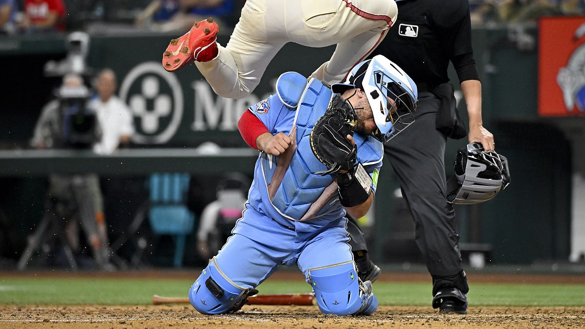 Toronto Blue Jays catcher Alejandro Kirk braces on his knees as Texas Rangers left fielder Wyatt Langford leaps over him at the plate.