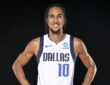 Dallas Mavericks guard Zaccharie Risacher smiles with his hands on his hips in a white home jersey against a black backdrop.