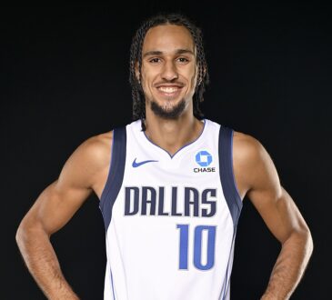 Dallas Mavericks guard Zaccharie Risacher smiles with his hands on his hips in a white home jersey against a black backdrop.