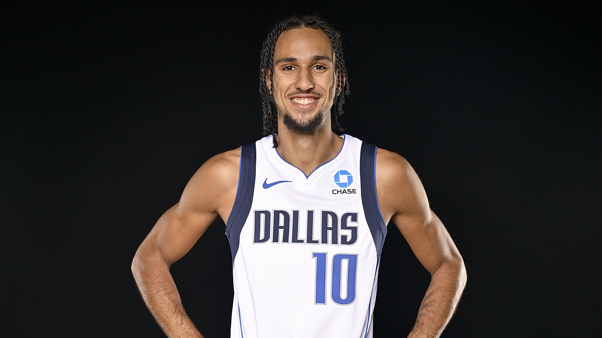 Dallas Mavericks guard Zaccharie Risacher smiles with his hands on his hips in a white home jersey against a black backdrop.