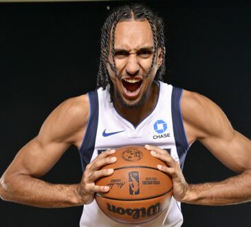 Dallas Mavericks forward Zaccharie Risacher roars toward the camera while gripping a basketball against a black backdrop.