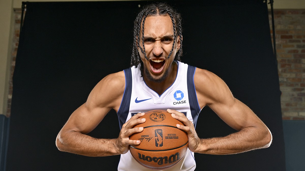 Dallas Mavericks forward Zaccharie Risacher roars toward the camera while gripping a basketball against a black backdrop.