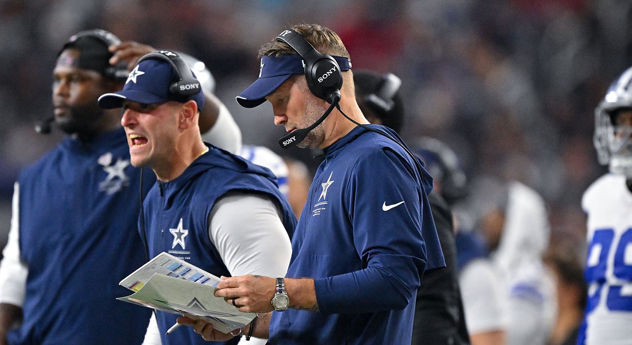 Cowboys head coach Brian Schottenheimer works the sideline in a headset alongside assistant coaches holding a play sheet.