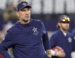 Cowboys head coach Brian Schottenheimer, wearing a team visor and pullover, watches warmups with his hands at his waist.