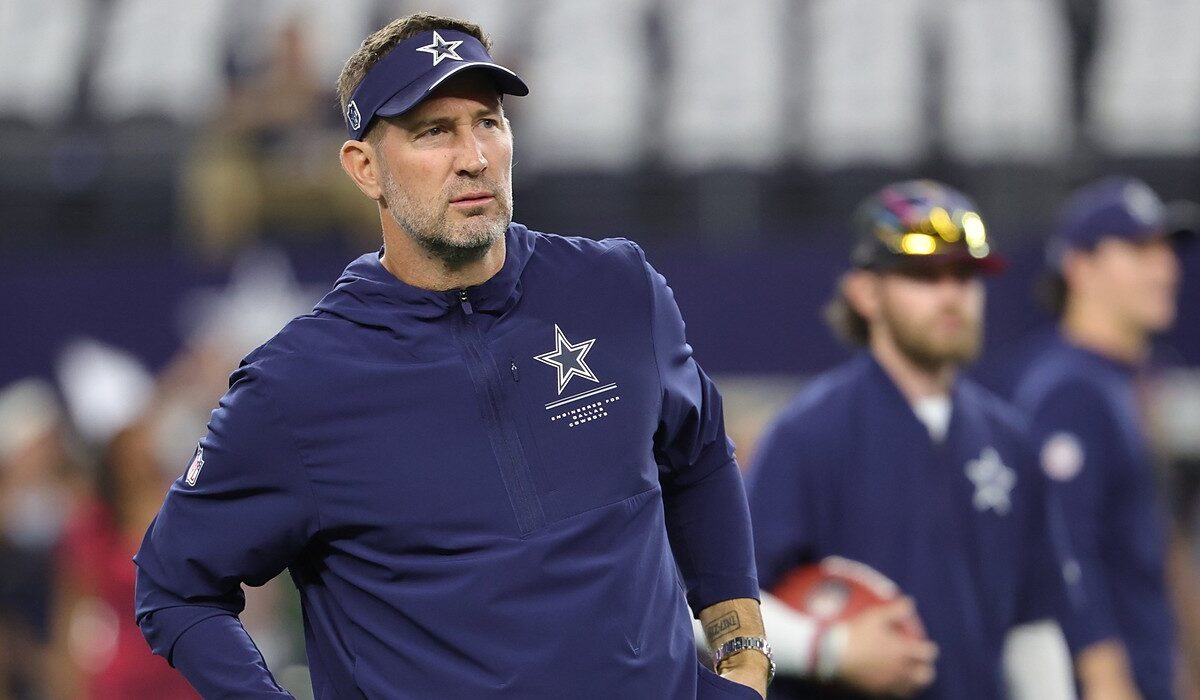 Cowboys head coach Brian Schottenheimer, wearing a team visor and pullover, watches warmups with his hands at his waist.