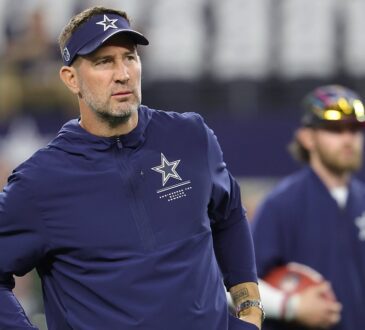 Cowboys head coach Brian Schottenheimer, wearing a team visor and pullover, watches warmups with his hands at his waist.