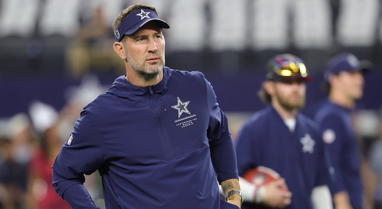 Cowboys head coach Brian Schottenheimer, wearing a team visor and pullover, watches warmups with his hands at his waist.