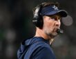 Dallas Cowboys head coach Brian Schottenheimer, wearing a headset and team visor, gazes upfield from the sideline.