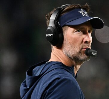Dallas Cowboys head coach Brian Schottenheimer, wearing a headset and team visor, gazes upfield from the sideline.