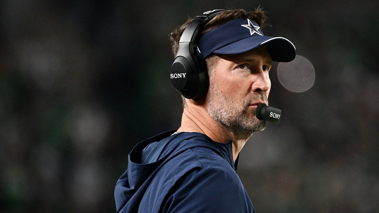 Dallas Cowboys head coach Brian Schottenheimer, wearing a headset and team visor, gazes upfield from the sideline.
