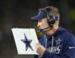 Dallas Cowboys head coach Brian Schottenheimer holds a play sheet while wearing a headset on the sideline.