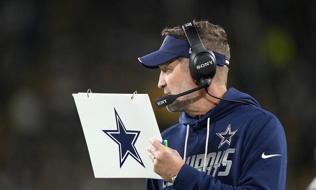 Dallas Cowboys head coach Brian Schottenheimer holds a play sheet while wearing a headset on the sideline.