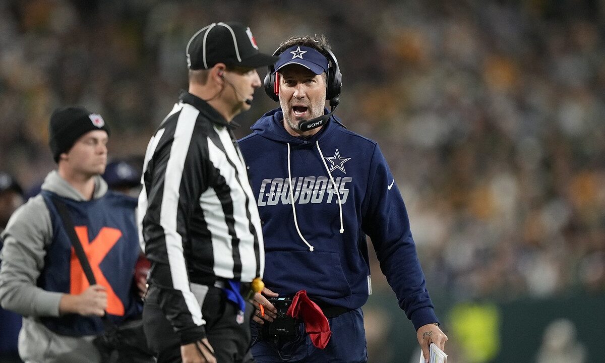 Dallas Cowboys head coach Brian Schottenheimer speaks to an official on the sideline while holding a red flag.
