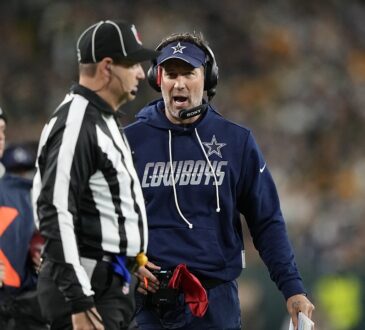 Dallas Cowboys head coach Brian Schottenheimer speaks to an official on the sideline while holding a red flag.