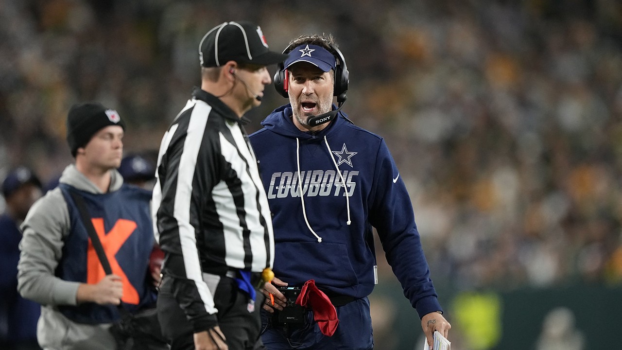 Dallas Cowboys head coach Brian Schottenheimer speaks to an official on the sideline while holding a red flag.