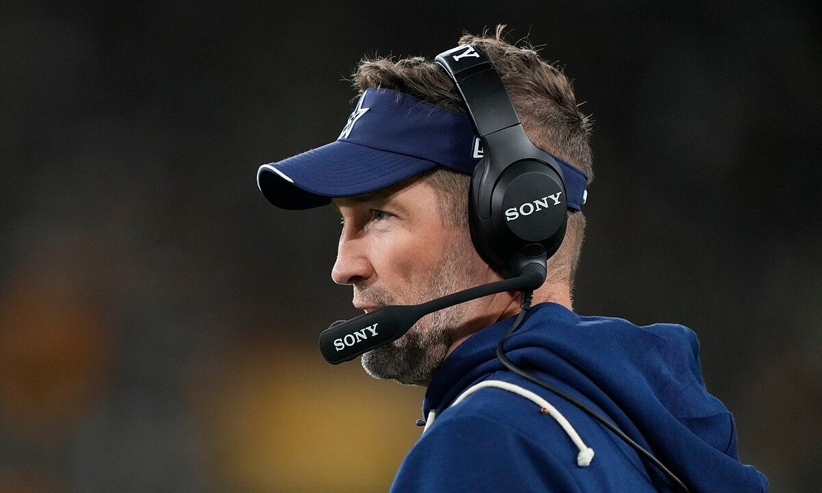 Dallas Cowboys head coach Brian Schottenheimer looks out at the field in profile, wearing a headset and team visor.