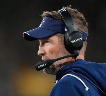 Dallas Cowboys head coach Brian Schottenheimer looks out at the field in profile, wearing a headset and team visor.