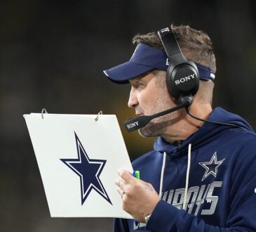 Dallas Cowboys head coach Brian Schottenheimer holds a play sheet while wearing a headset on the sideline.