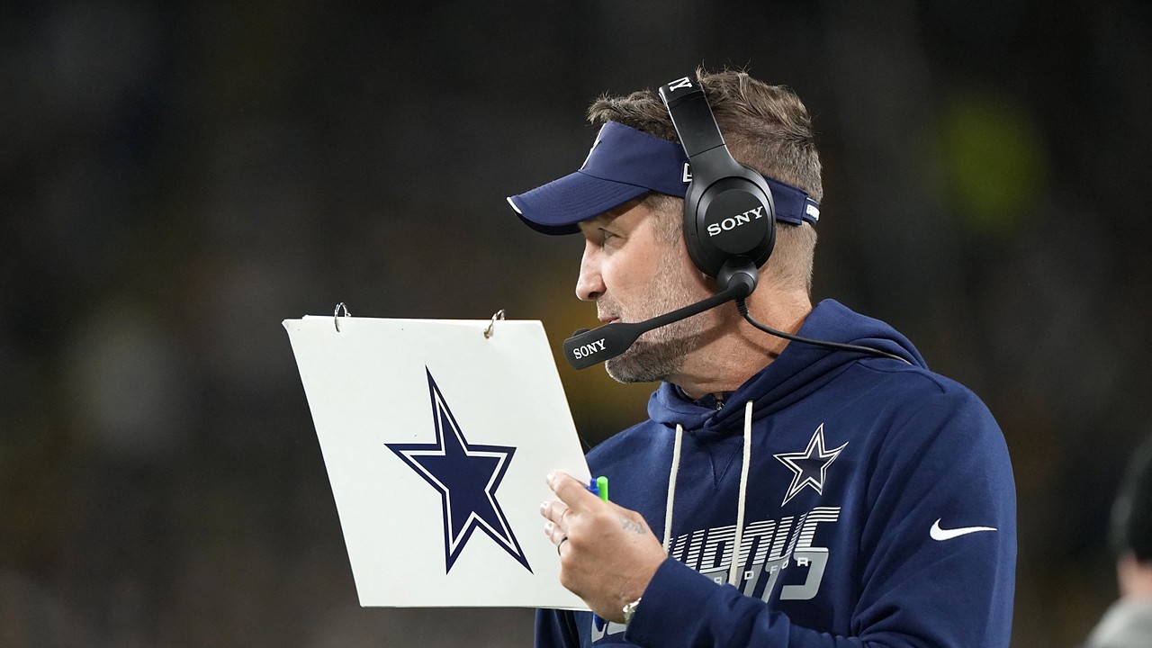 Dallas Cowboys head coach Brian Schottenheimer holds a play sheet while wearing a headset on the sideline.